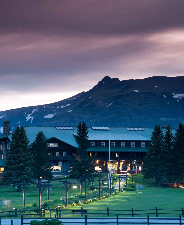 The warm glow of the windows of Glacier Park Lodge at dusk with dark blue mountains looming in the background.
